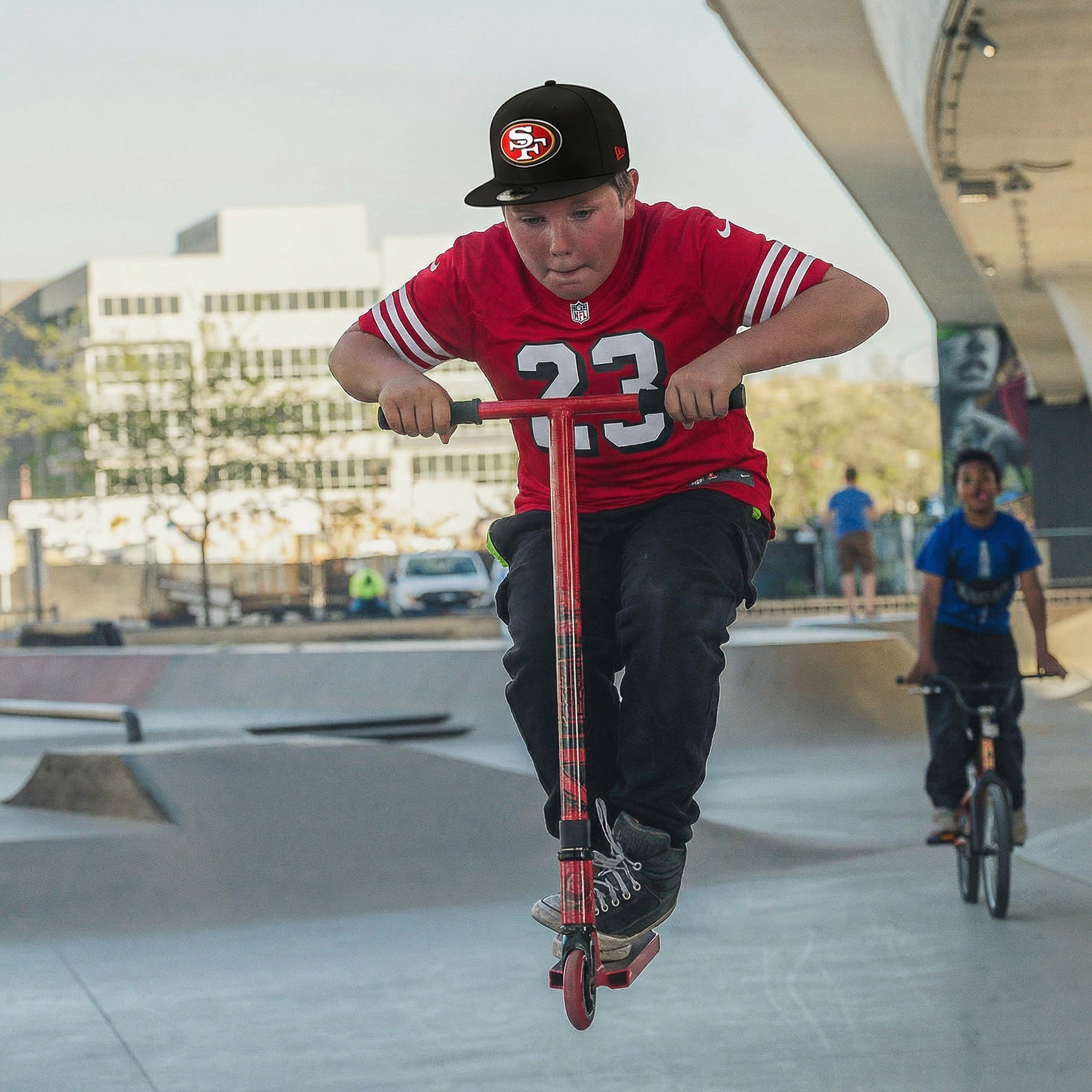 Person riding a scooter at a skate park with another person on a bicycle in the background.